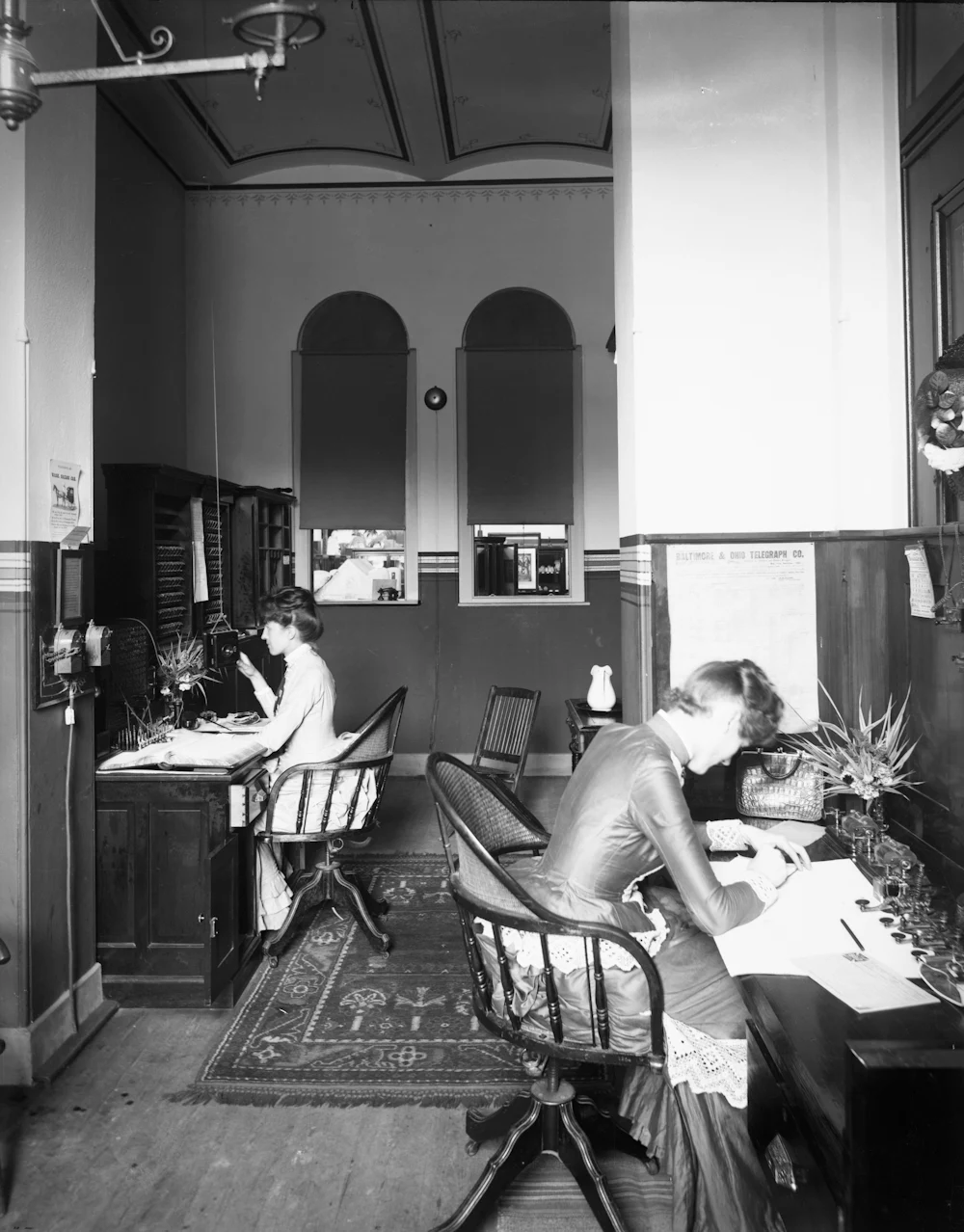 A telephone and telegraph office around 1900: two operators at desks, one seated at a small switchboard